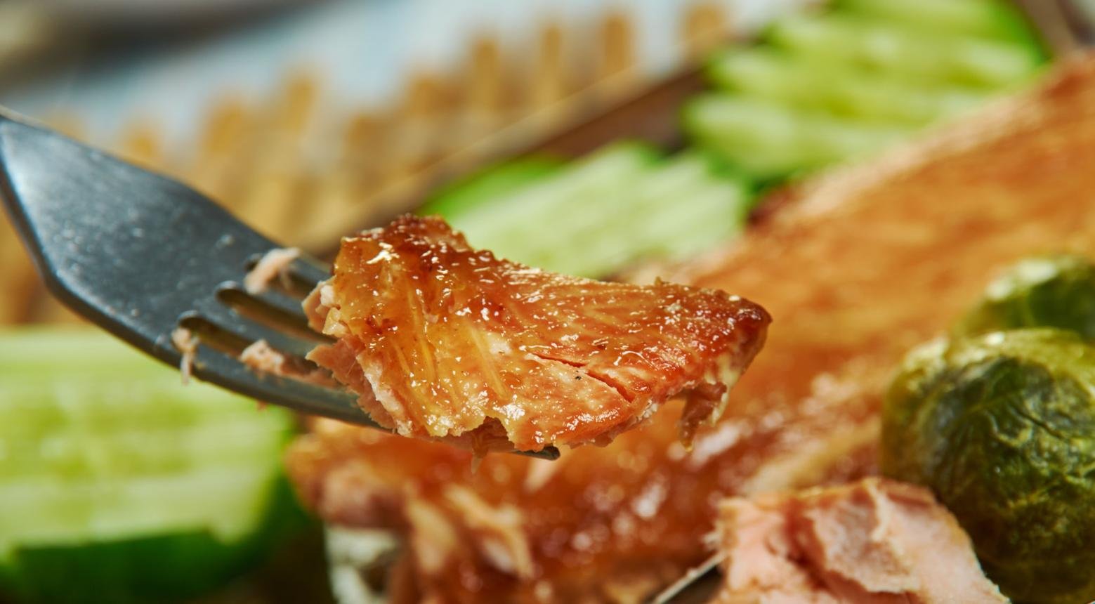 Fork with a glossy, roasted meat bite, crispy crust, green veggies blurred in background.