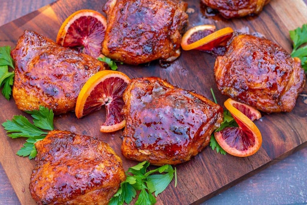 Glazed roast chicken thighs on a wooden board, garnished with orange slices and parsley.