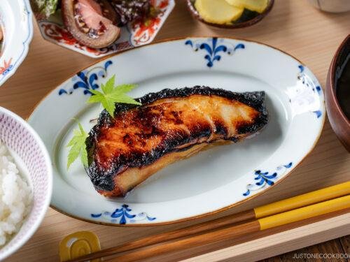 Grilled salmon fillet on a white plate with blue trim, garnished with a green leaf, with a bowl of rice and chopsticks nearby.