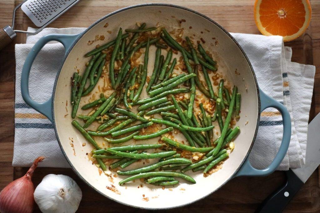Top-down view of a blue-handled skillet filled with sautéed green beans and browned onion/garlic bits on a wooden counter.