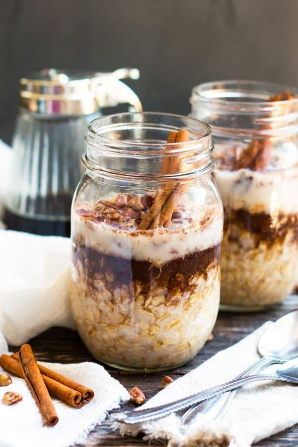 Two mason jars of overnight oats with yogurt, chocolate, and cinnamon sticks on a wooden table.