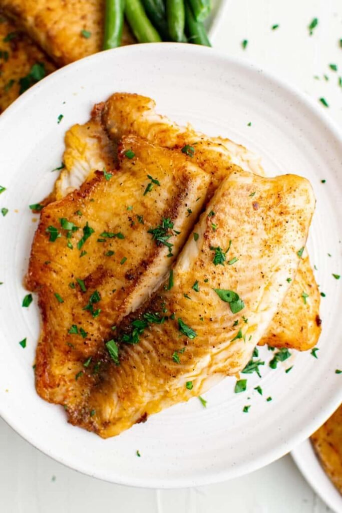 Two pan-seared fish fillets with a golden crust, sprinkled with parsley on a white plate with green beans in the background