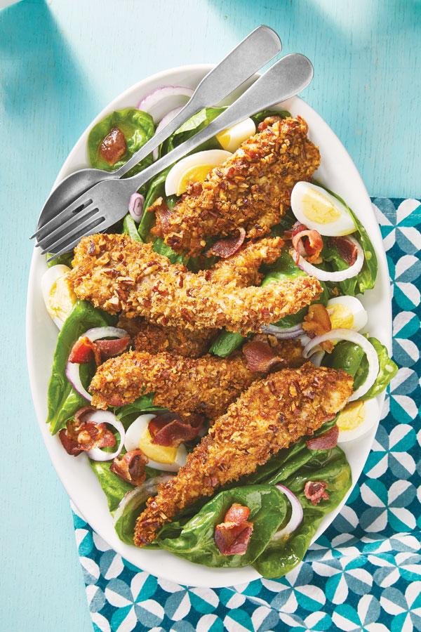 Plate of crispy fried chicken tenders on a bed of greens with hard-boiled egg wedges, red onions, and bacon bits.