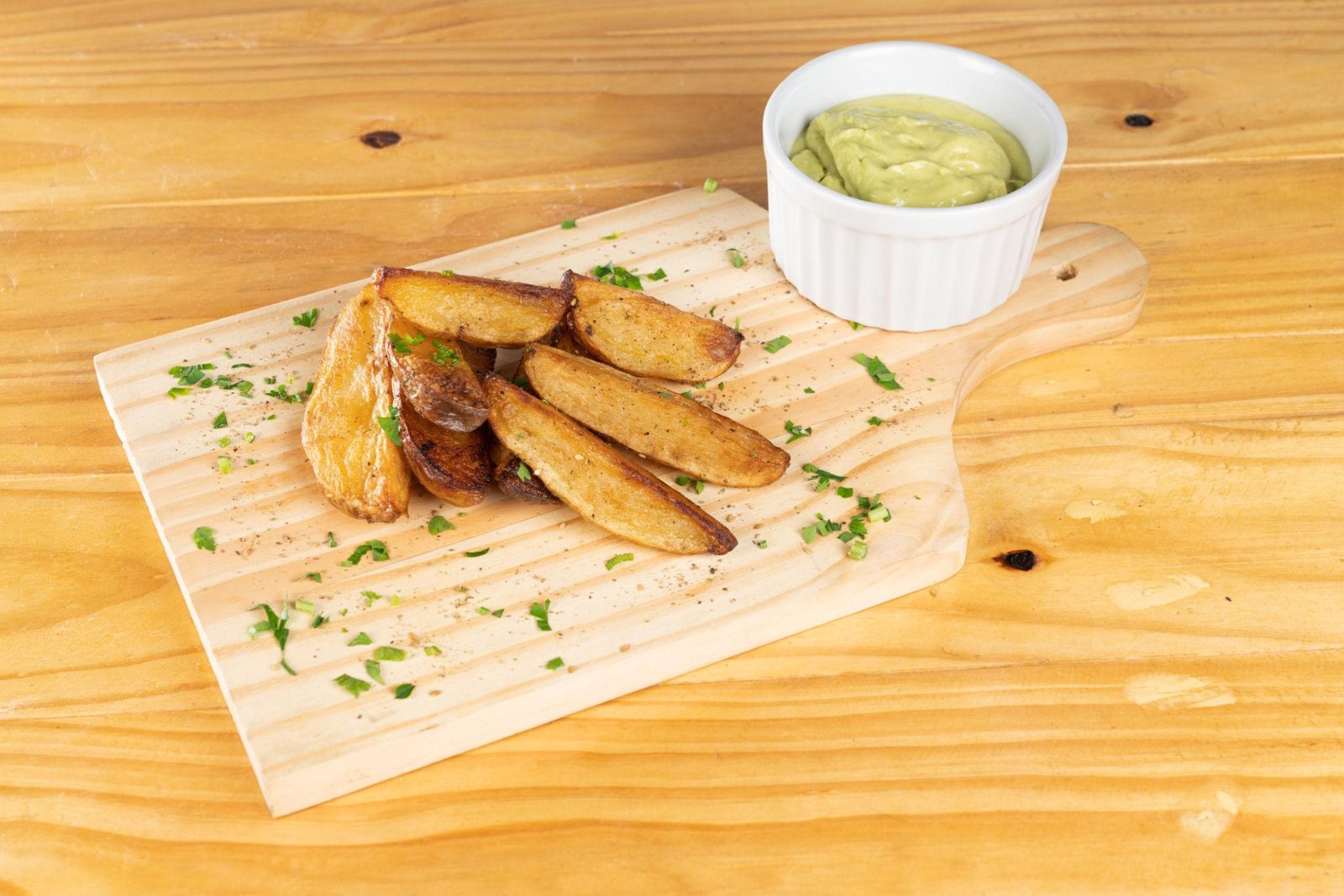 Crispy potato wedges on a wooden board with a small white ramekin of green dipping sauce beside them.