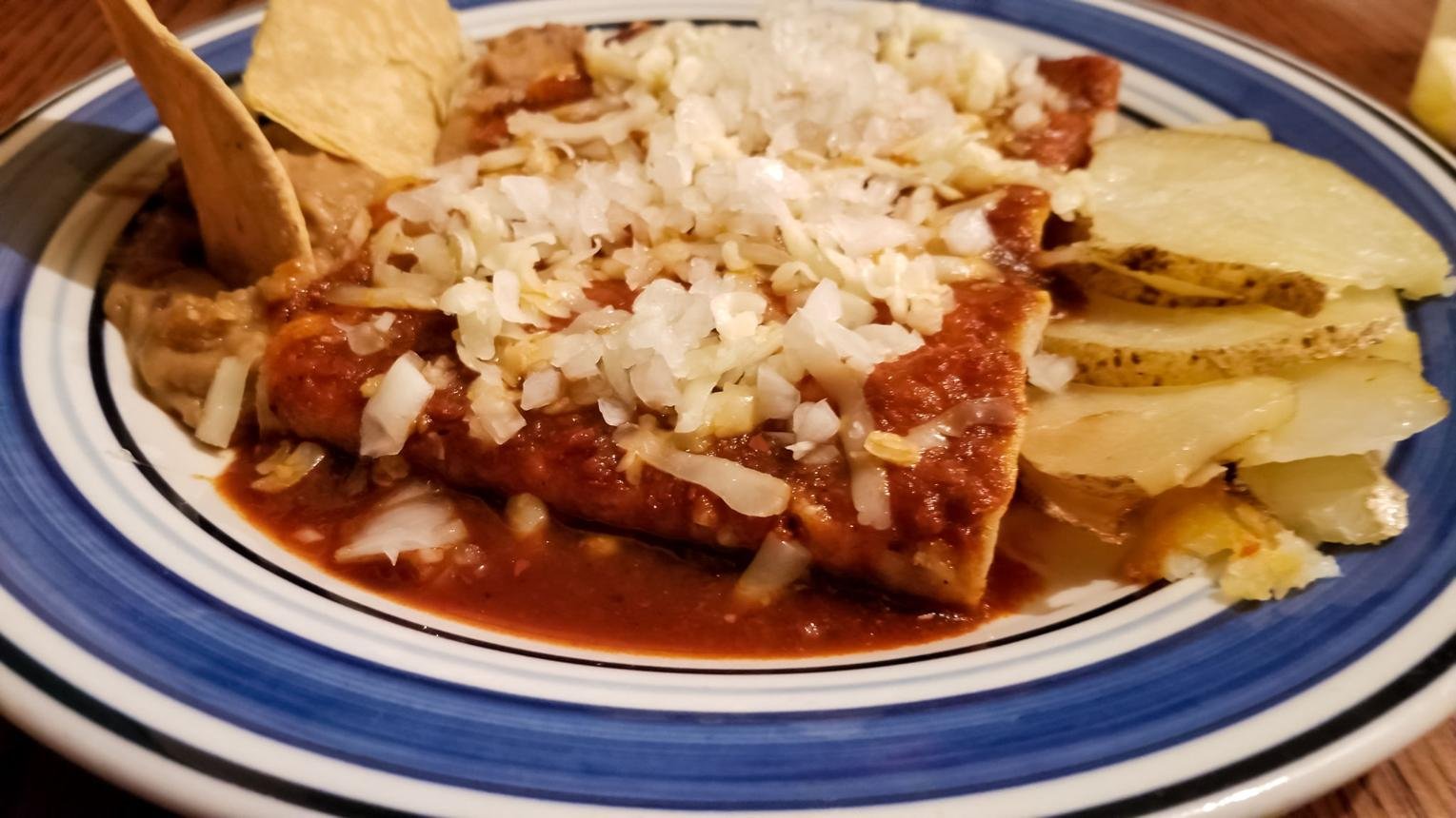 Enchiladas in red sauce topped with chopped onions, served with potato wedges on a blue-rimmed plate.