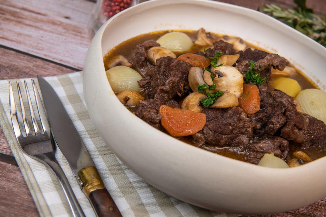 Beef stew with potatoes, onions, carrots, and mushrooms in a white bowl on a wooden table.