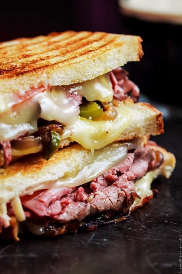 Stacked grilled roast beef sandwich with melted cheese and sautéed peppers between toasted slices on a dark surface.