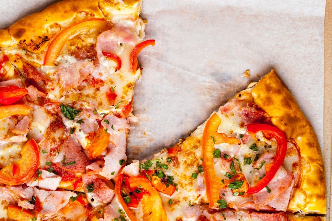 Two slices of loaded pizza on parchment with ham, red peppers, and herbs showing melted cheese and a golden crust