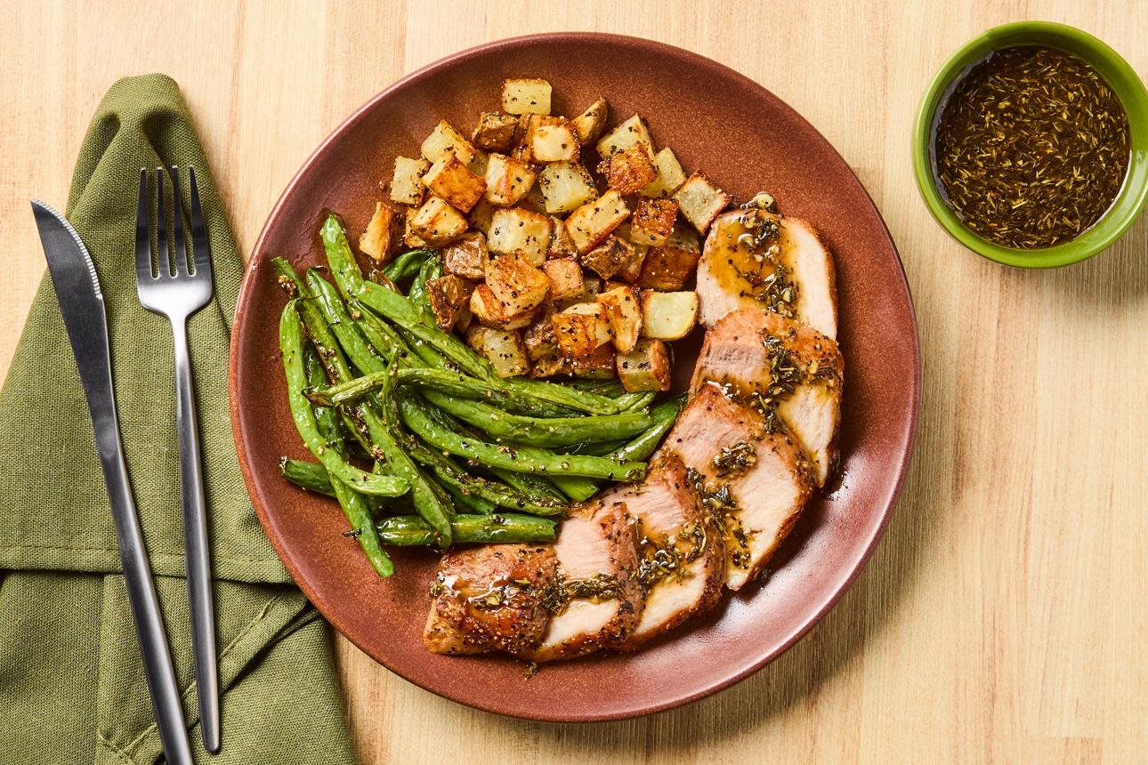 Sliced herb-crusted pork tenderloin with roasted potatoes and green beans on a brown plate, plus a side of herb dressing in a green cup.