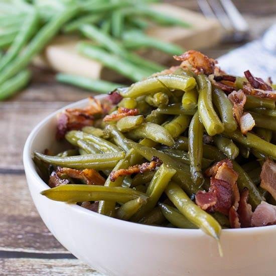Sautéed green beans with crispy bacon in a white bowl on a wooden table.