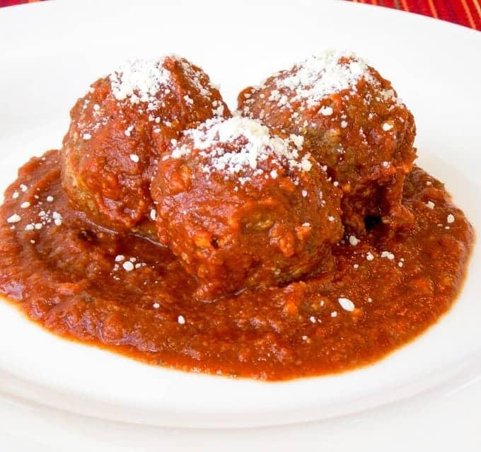 Three meatballs in a chunky tomato sauce, topped with grated cheese on a white plate.