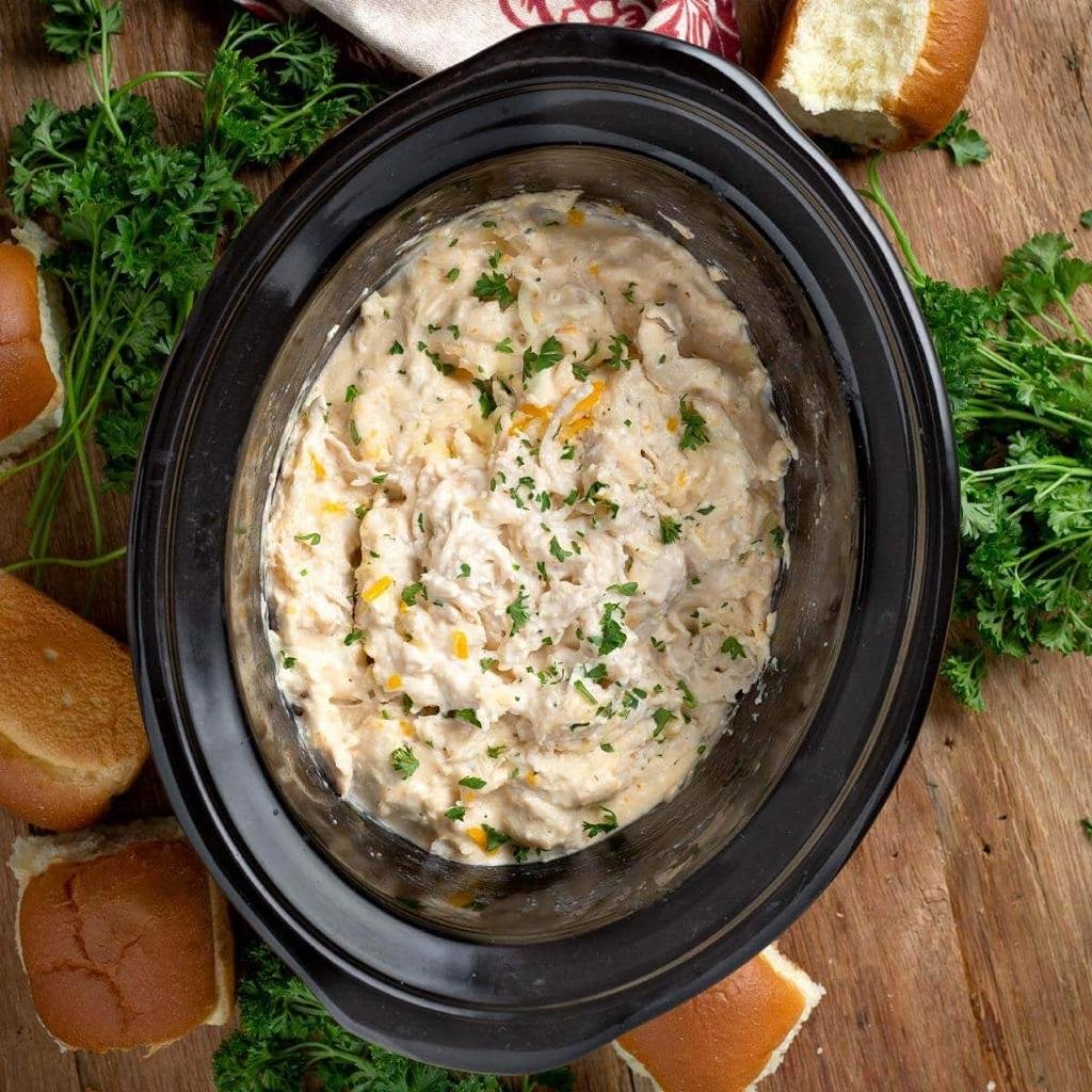 Creamy dip in a black crockpot, garnished with chopped parsley, with crusty bread and herbs around on a wooden table.
