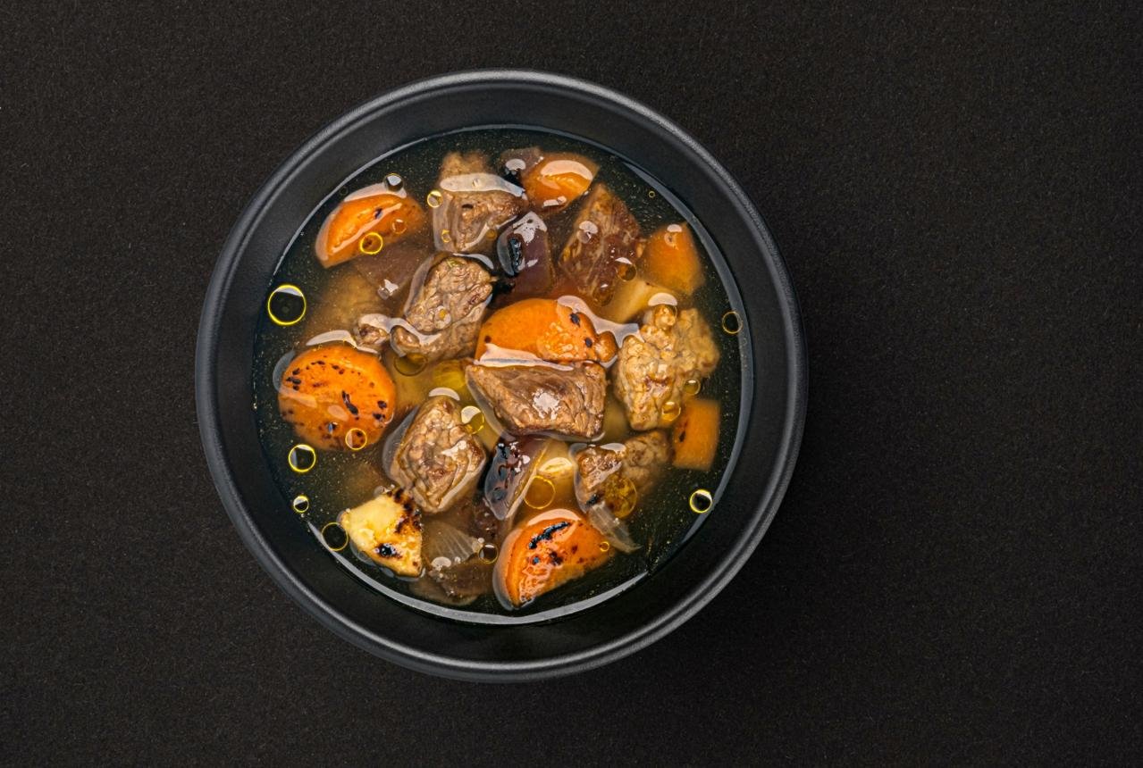 Black bowl containing beef stew with chunks of meat and orange carrots in a clear broth on a dark surface
