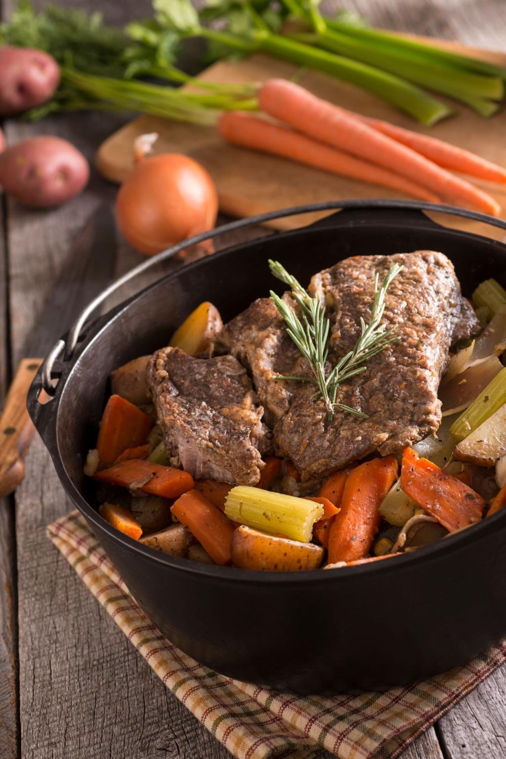 Beef pot roast with carrots, potatoes, and celery in a dark Dutch oven, topped with a sprig of rosemary on a rustic table