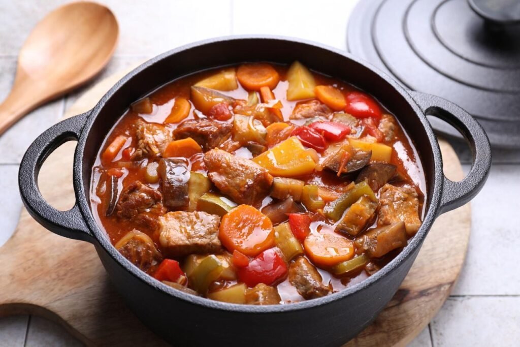 Beef stew with chunks of carrot, potato, and peppers in a dark cast-iron pot on a wooden board