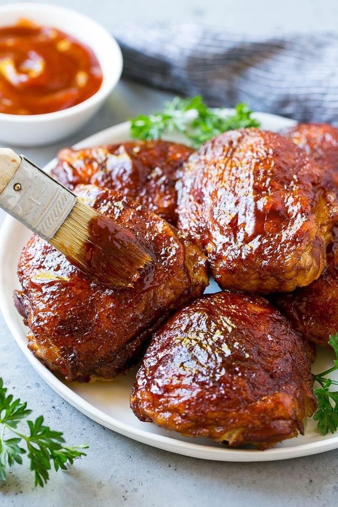 Plate of glossy barbecue chicken thighs being brushed with sauce, parsley garnish, and a bowl of dipping sauce.