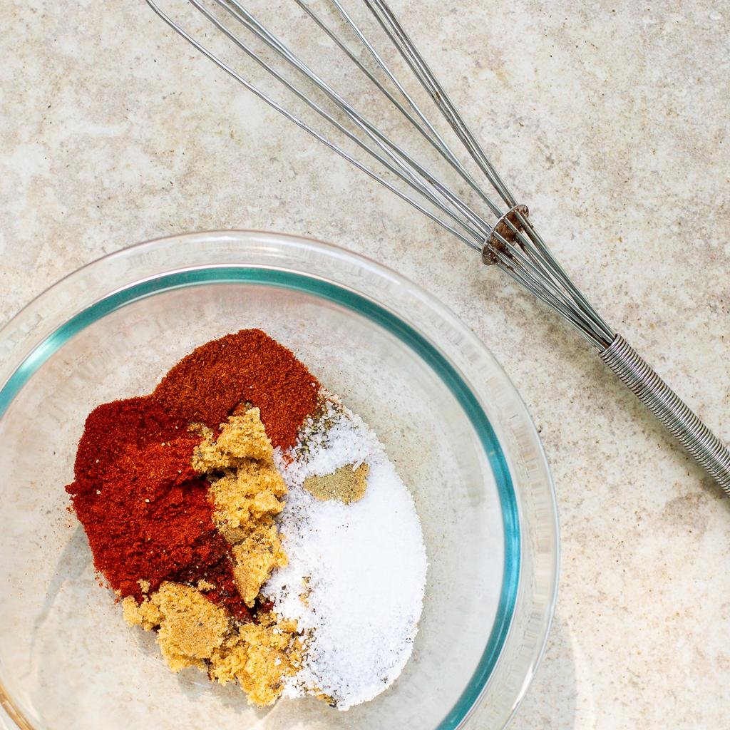 Bowl with red chili powder, brown sugar, white sugar, and salt beside a metal whisk on a beige countertop.