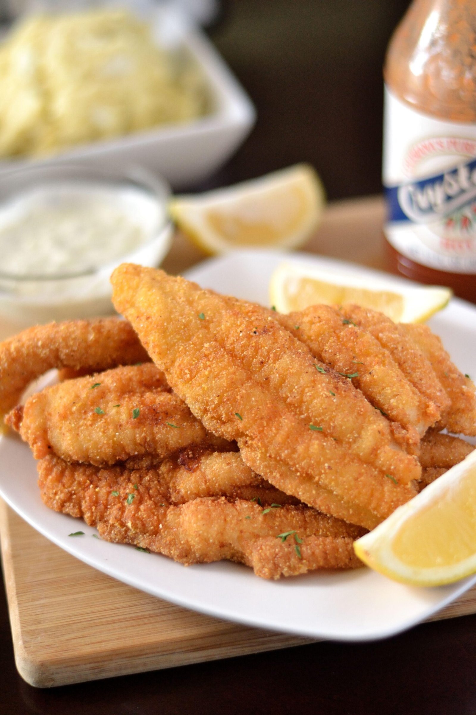 Crispy breaded fried fish fillets piled on a white plate with lemon wedges and a small bowl of dipping sauce in the background