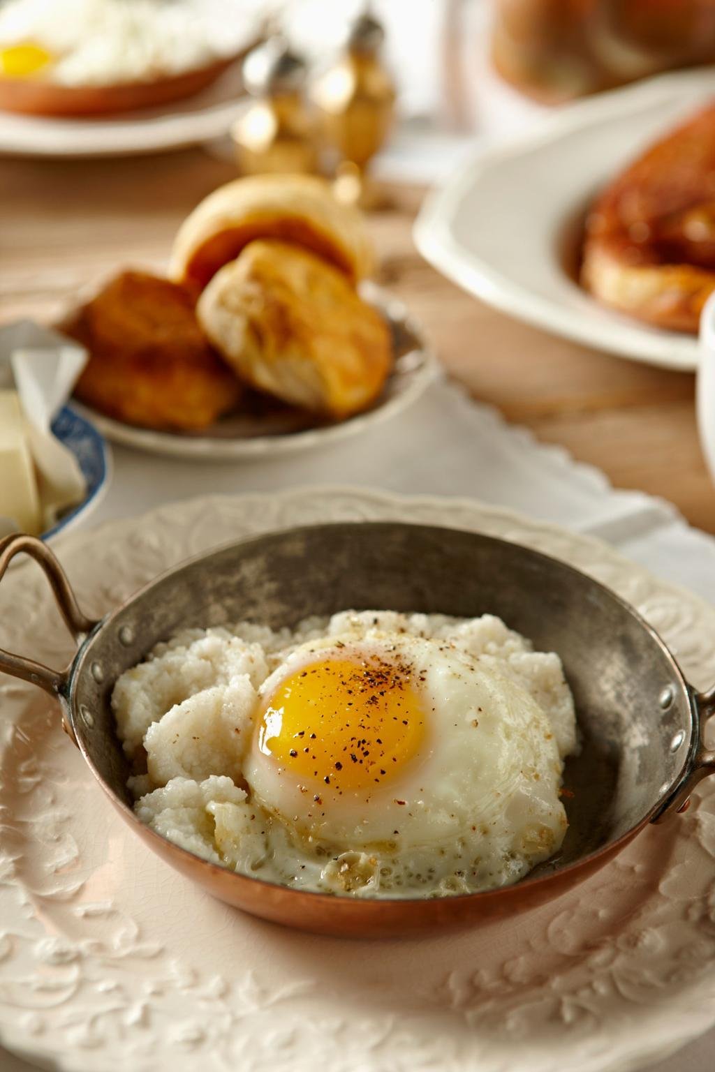 Sunny-side-up egg over creamy mashed potatoes in a copper skillet, sprinkled with pepper, on a breakfast table with bread rolls in the background.