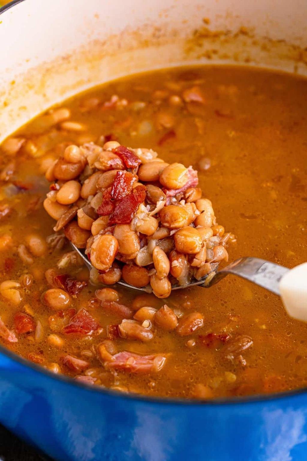 A spoonful of baked beans with bits of bacon held over a simmering pot of bean chili in a blue pot.