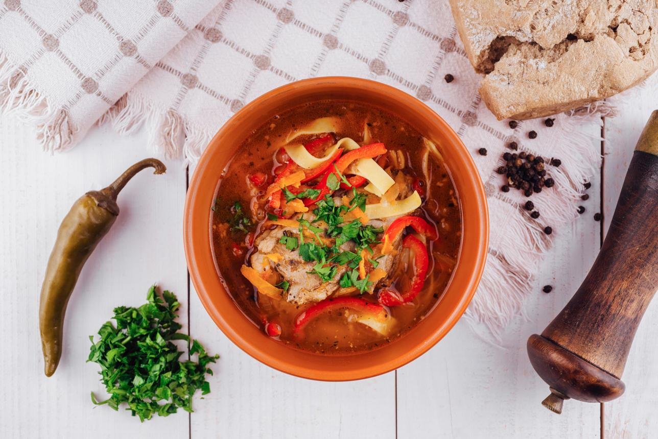 A terracotta bowl of hearty meat soup topped with herbs, red peppers, and cheese shreds, on a white wooden table with chili pepper, chopped parsley, bread, and a wooden mortar nearby.