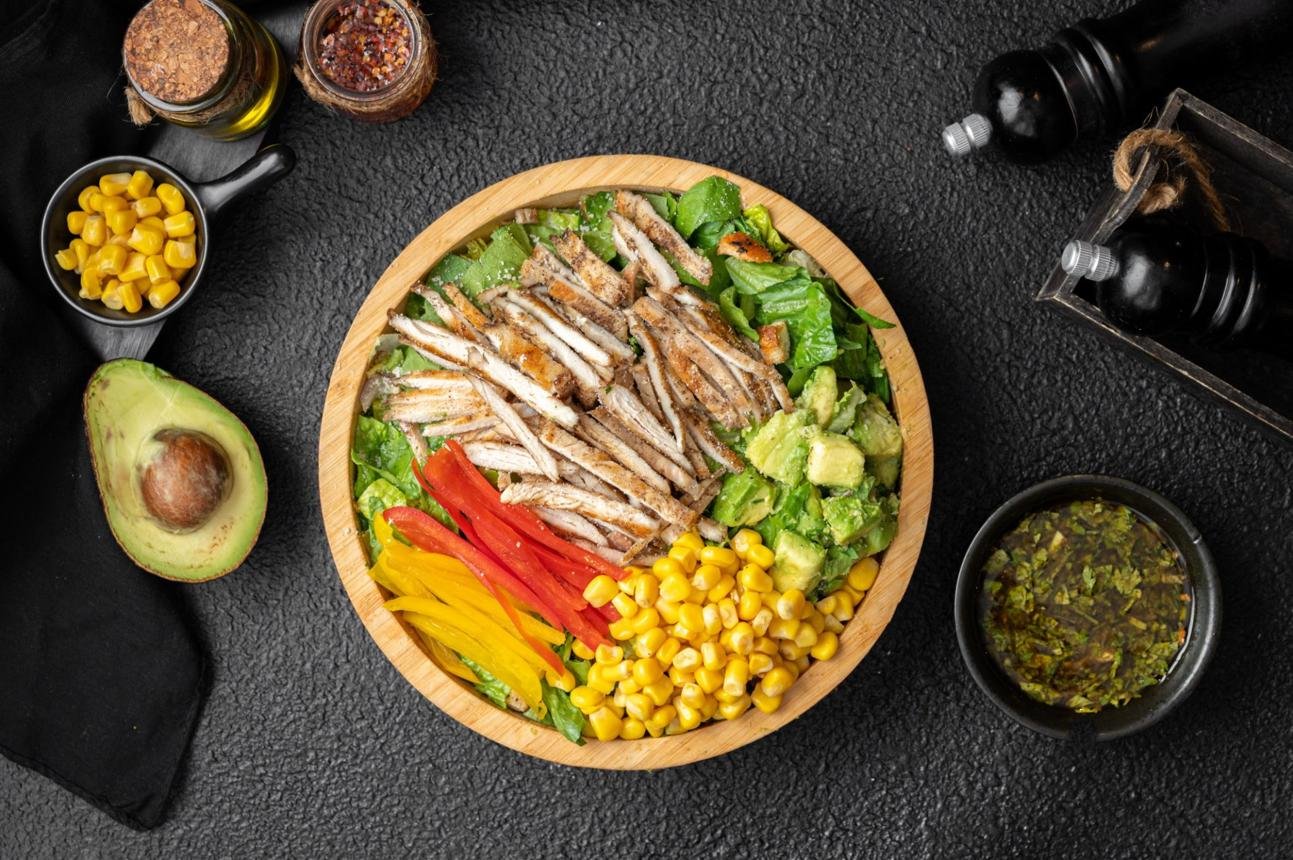 Bowl of mixed salad with sliced grilled fish, avocado, corn, red and yellow peppers on a wooden plate.