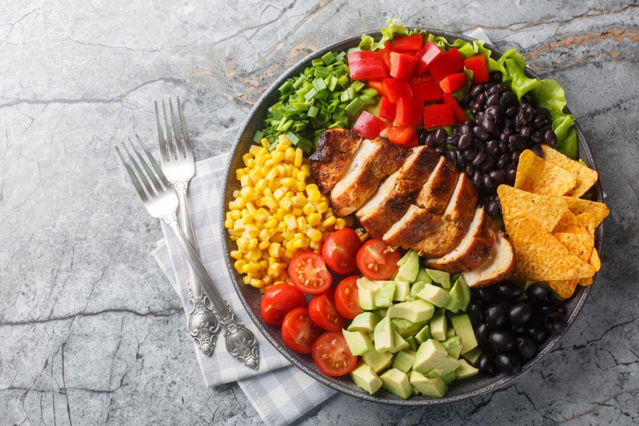 Colorful plated salad with grilled chicken slices, corn, tomatoes, avocado, black beans, bell peppers, greens, and tortilla chips on a round platter