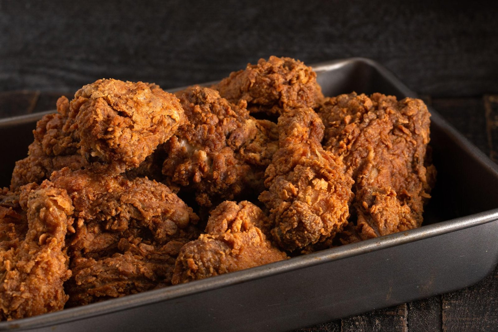Pile of crispy fried chicken pieces in a dark metal baking tray on a wooden surface, golden-brown and crunchy.