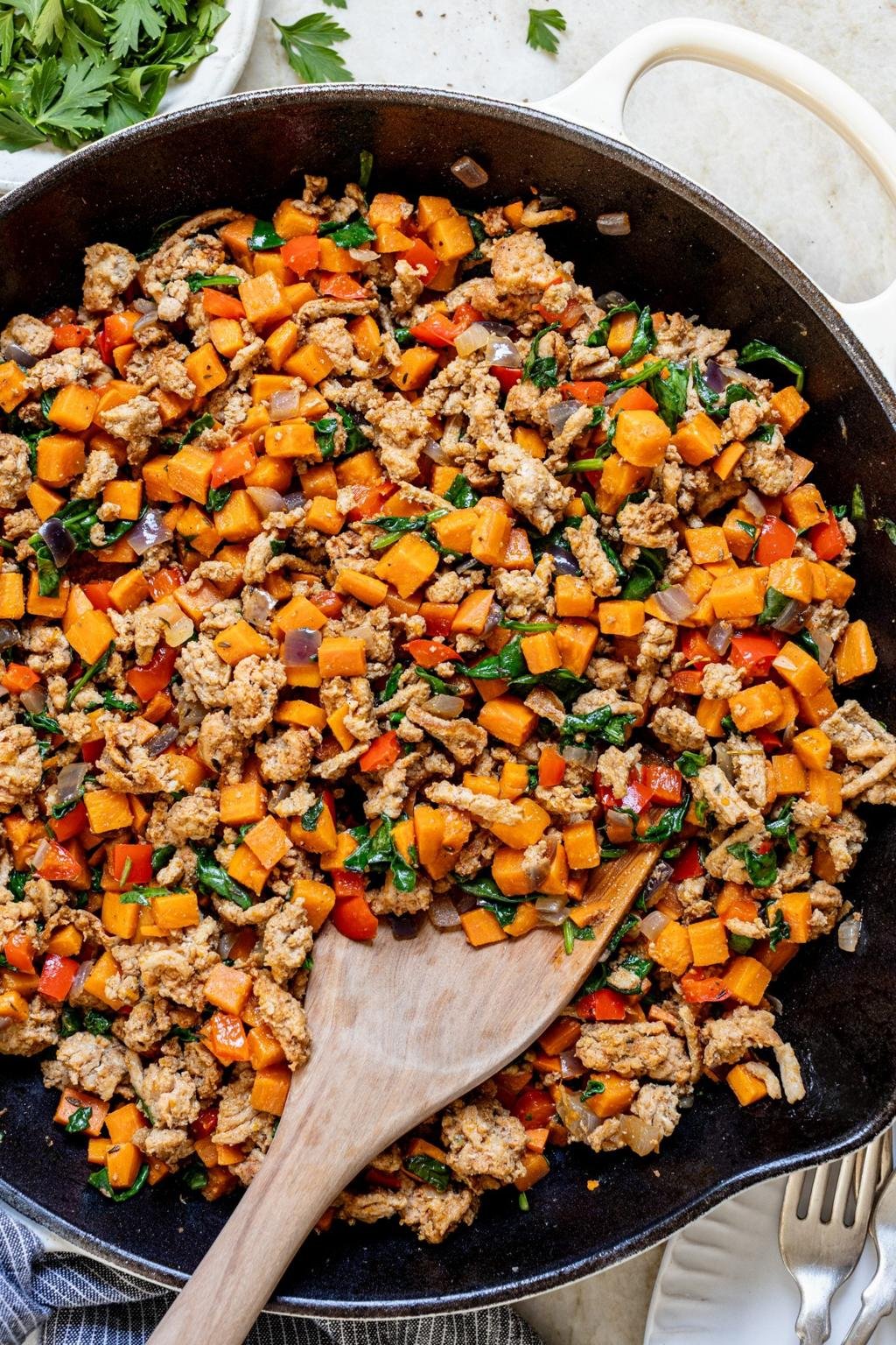 Skillet hash with browned ground meat, diced sweet potatoes, red peppers, onions, and greens being stirred with a wooden spatula.