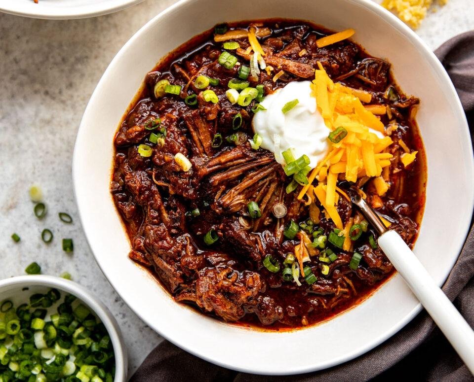 Bowl of braised beef in a dark chili-like sauce, topped with sour cream, shredded cheddar, and chopped green onions.