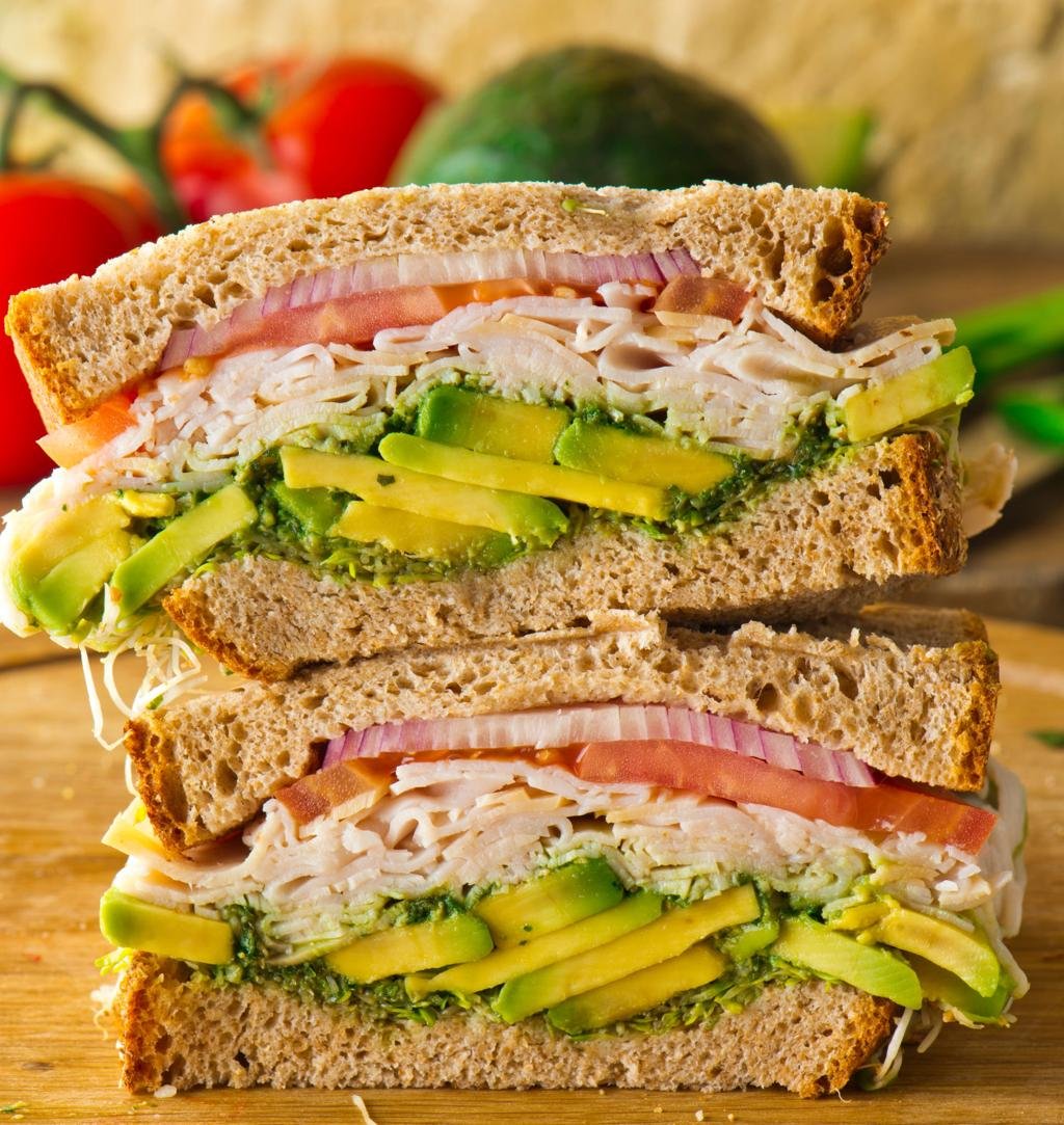 Cross-section of stacked whole-grain sandwiches filled with turkey, avocado, tomato, and greens on a wooden board.
