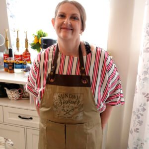 Smiling woman in a striped shirt wearing a brown apron in a kitchen with bottles on the counter behind her.