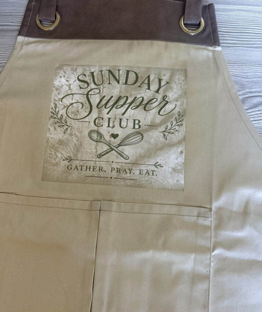 Beige apron with brown leather top and brass rings, showing a 'Sunday Supper Club' patch with a whisk and spoon graphic and the words 'Gather. Pray. Eat.'